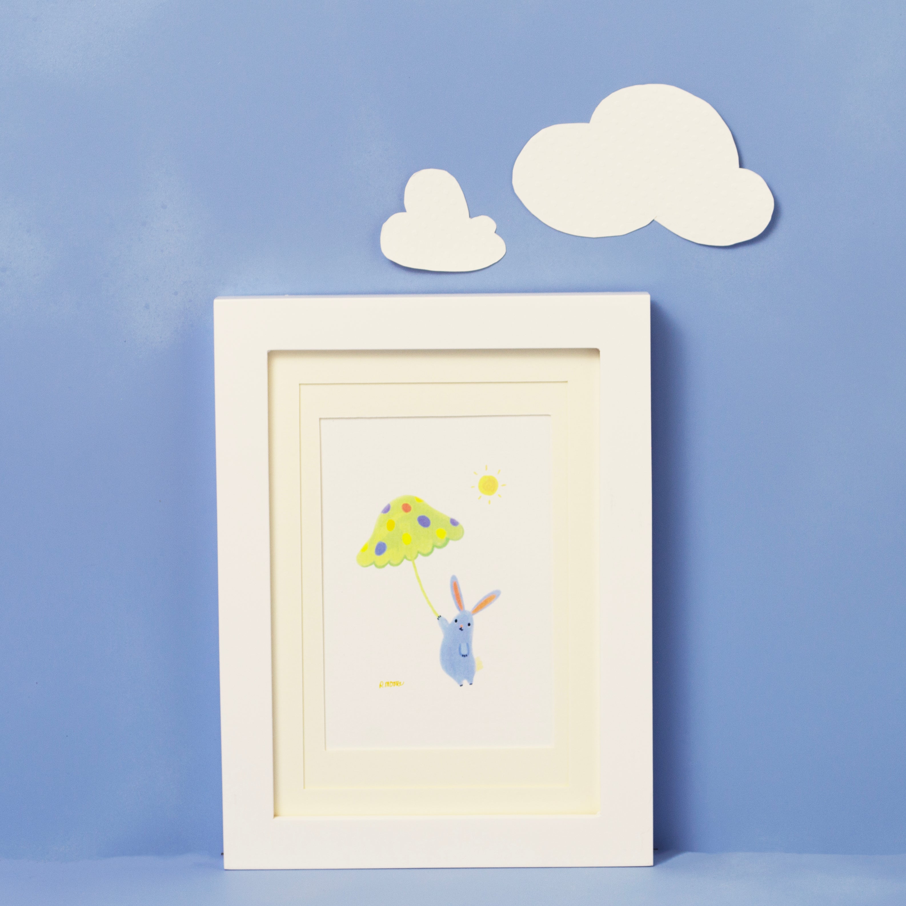 Bunnies with Ideas framed print of a hand illustrated blue rabbit holding a toadstool umbrella against a white background. The framed print is shot against a blue background and two white clouds aove the framed print. The frame is white, and the matting is ivory