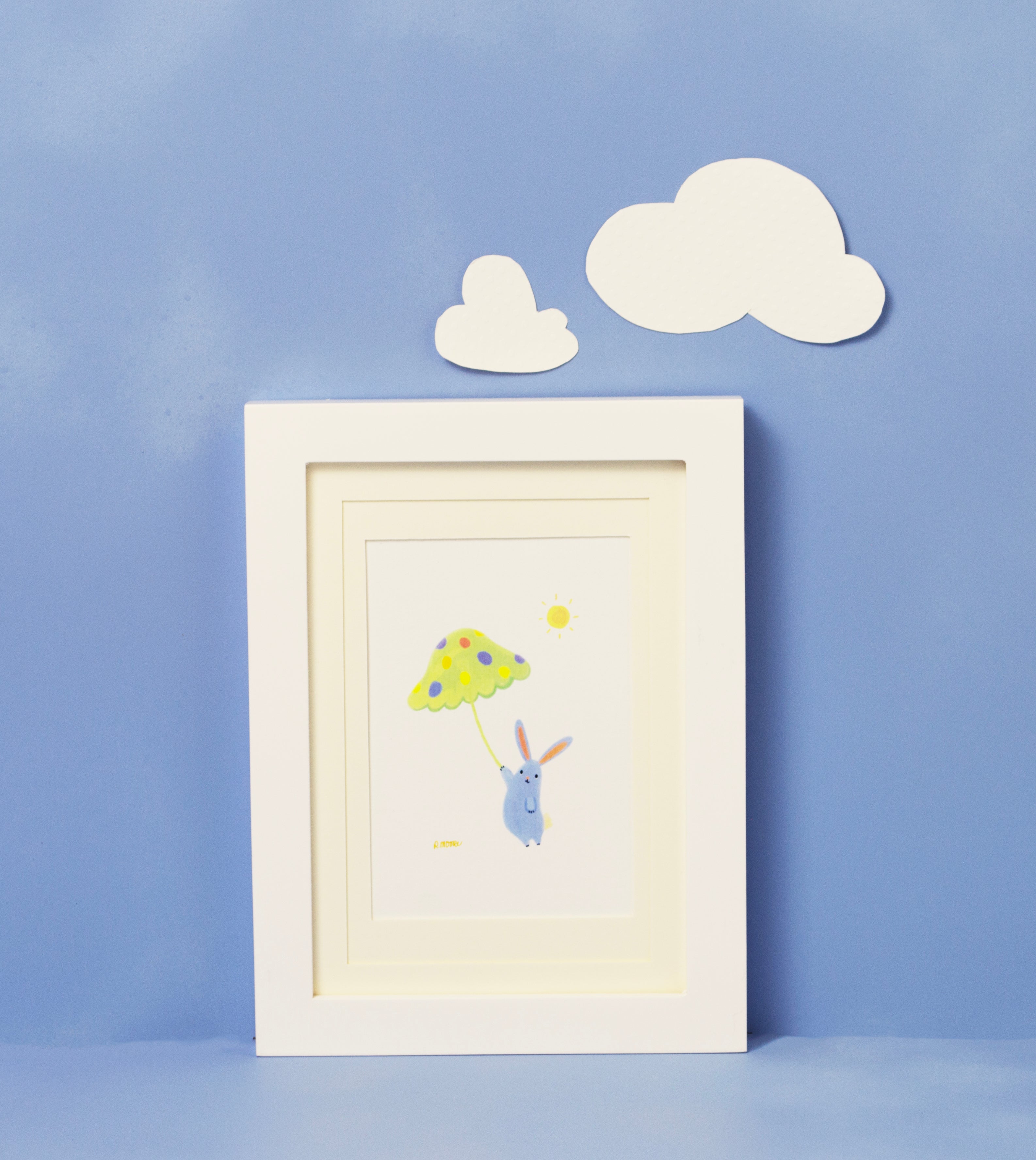 Bunnies with Ideas framed print of a hand illustrated blue rabbit holding a toadstool umbrella against a white background. The framed print is shot against a blue background and two white clouds aove the framed print. The frame is white, and the matting is ivory